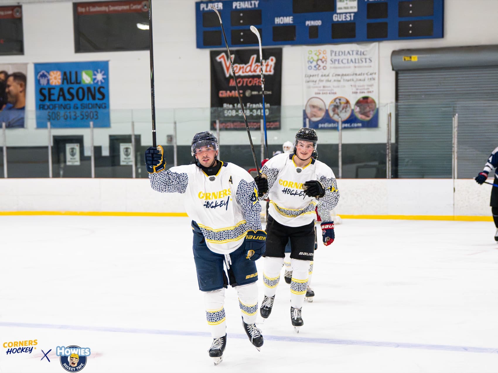 Two Corners Hockey players in white jerseys skating back up the ice, one with a fist raised after a goal
